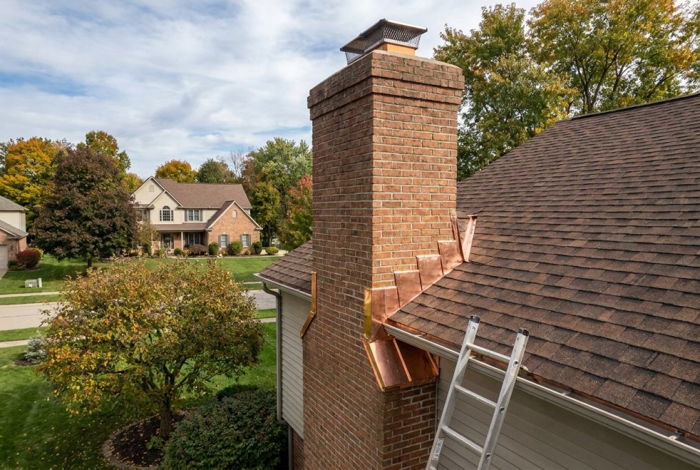 Copper chimney flashing being installed on a residential roof with a ladder in a suburban neighborhood